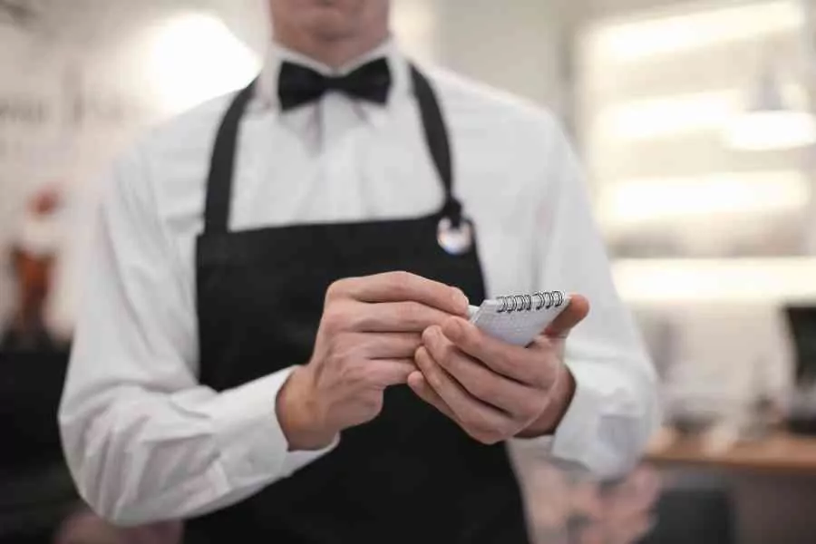 A Waiter In A White Shirt, Black Vest, And Bow Tie Holds A Notepad And Pen, Ready To Take An Order In A Restaurant Setting.