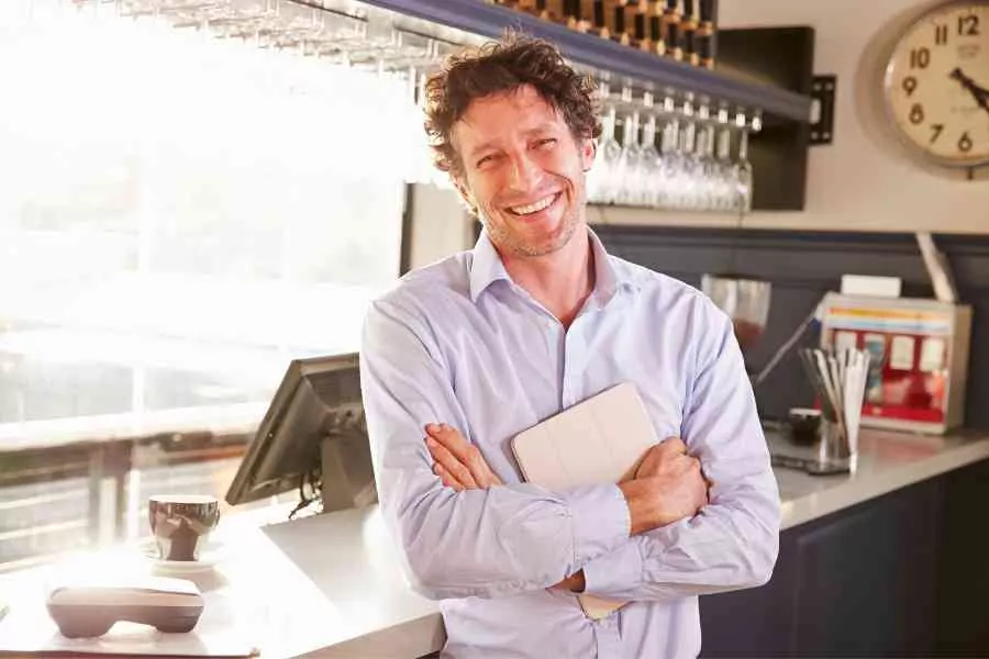 A Smiling Man In A Blue Shirt Standing With Crossed Arms In A Sunlit Bar, Holding A Tablet.