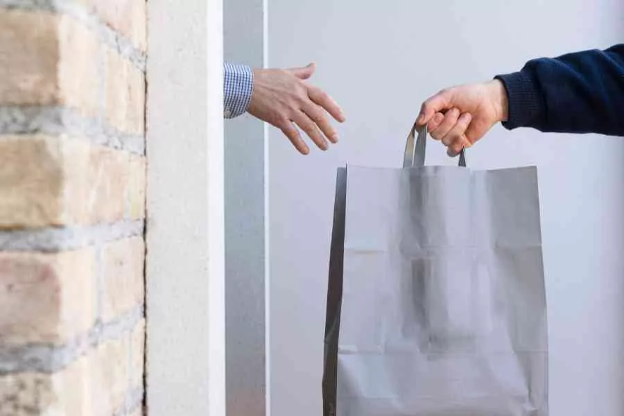 Two Hands Exchanging A Gray Shopping Bag Through A Doorway, Highlighting A Transaction Or Delivery.