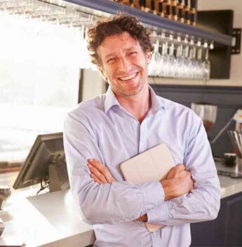 A Smiling Man In A Blue Shirt Standing With Crossed Arms In A Sunlit Bar, Holding A Tablet.