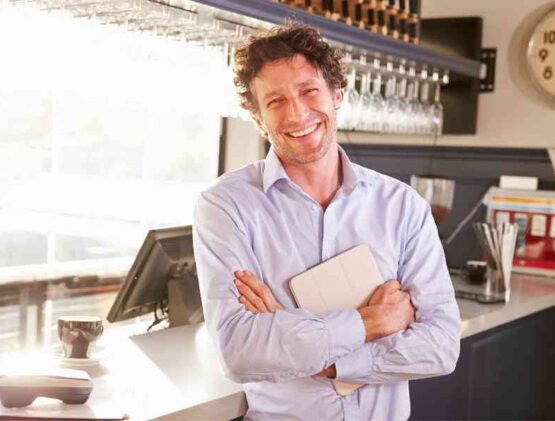 A Smiling Man In A Blue Shirt Standing With Crossed Arms In A Sunlit Bar, Holding A Tablet.