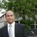 A Man In A Business Suit Smiling At The Camera, Standing Outdoors With Trees And An Auto Draft Building In The Background.