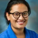 Portrait Of A Smiling Woman With Glasses, Wearing A Blue Shirt. She Has Long Dark Hair Pulled Back, Against A Blurred Blue Background. The Subject, Sanju Kunwar, Ph.d., Exudes