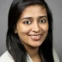 A Professional Headshot Of A Smiling Woman With Long Dark Hair, Wearing An Elegant White Blouse, Against A Gray Background.