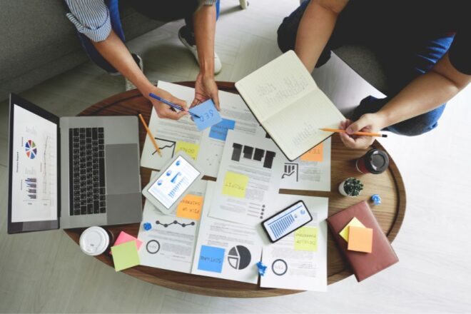 Overhead View Of A Business Meeting With Four People, Laptops, Documents, And Coffee On A Round Table.