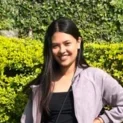 A Woman With Long Dark Hair Smiling, Wearing A Gray Jacket And Auto Draft Top, Standing In Front Of Lush Green Bushes In Sunlight.