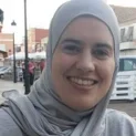 A Smiling Woman Wearing A Gray Hijab Sits Outdoors At A Café, With A Street And Parked Autos In The Background.