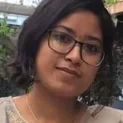 A Young Woman With Glasses, Wearing A Light Colored Blouse, Poses For An Auto Draft Photo In Front Of Blurred Green Foliage.