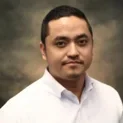 Portrait Of Sumit Pradhan, A Middle Aged Academic With A Faint Smile, Wearing A White Shirt, Set Against A Dark Mottled Background.