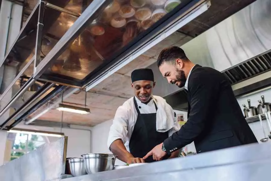 Two Men, One In Chef Attire And The Other In A Suit, Collaborating And Smiling In A Professional Kitchen.